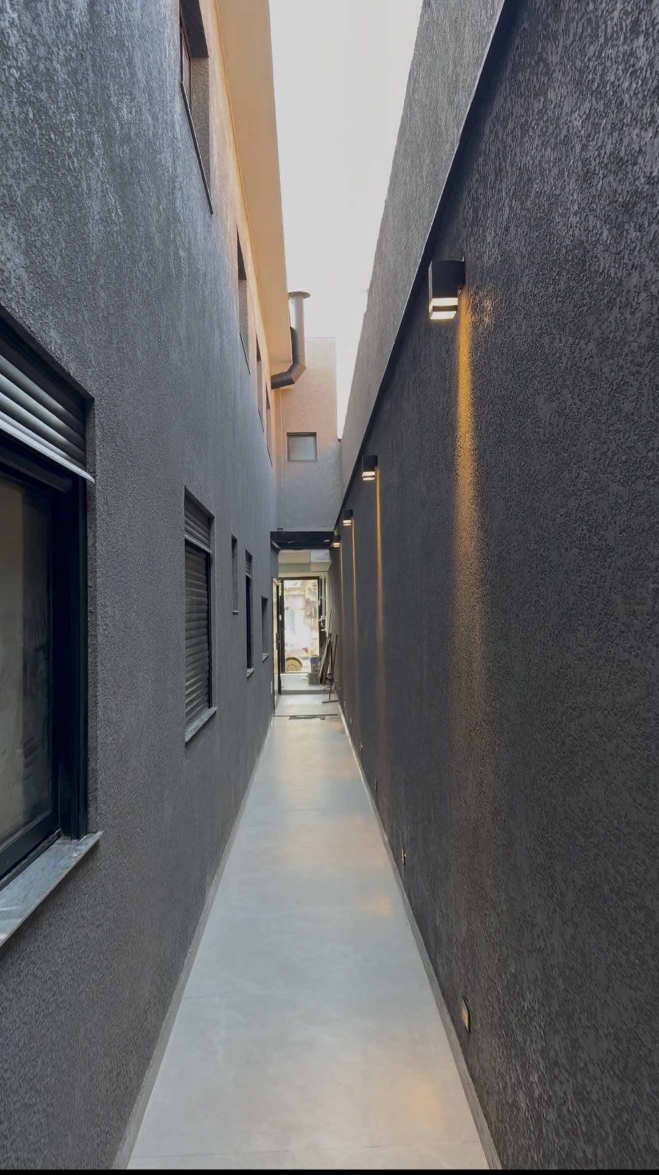 Obra da TM Soluções: corredor externo com textura projetada cinza e iluminação quente de arandelas